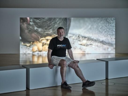 Kuvassa on Jorma Saarikko istumassa näyttelytilassa videoteoksen edessä. Hän on keski-ikäinen lyhythiuksinen mies, jolla on päällä musta t-paita, vaaleat shortsit ja mustat lenkkarit.